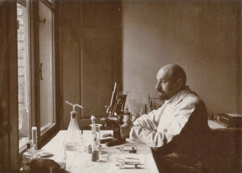 Profesor Adam Wrzosek siedzi przy stole. Patrzy w kierunku okna. Na stole stoi mikroskop.